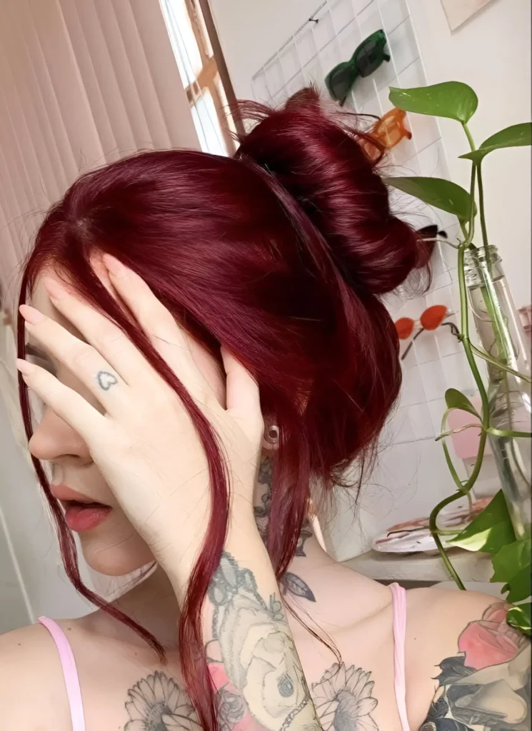 Person with bright burgundy hair, partially styled in a high, voluminous bun, with face partially obscured.