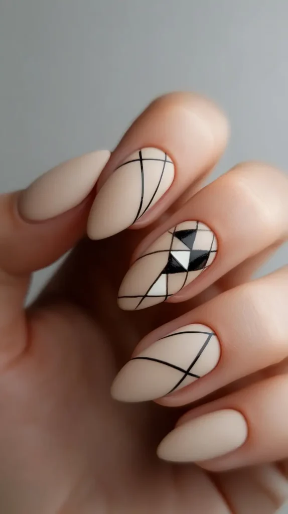 Close-up of a hand with matte nude, almond-shaped nails featuring minimalist black geometric line art; one nail has an intricate black and white mosaic pattern.