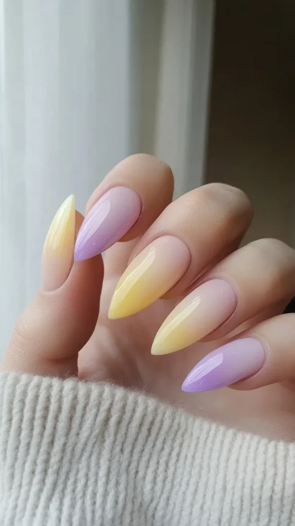 Close-up of long stiletto or almond nails with a sheer milky base fading into alternating pastel yellow and lavender ombre tips, resting on a knit sleeve.