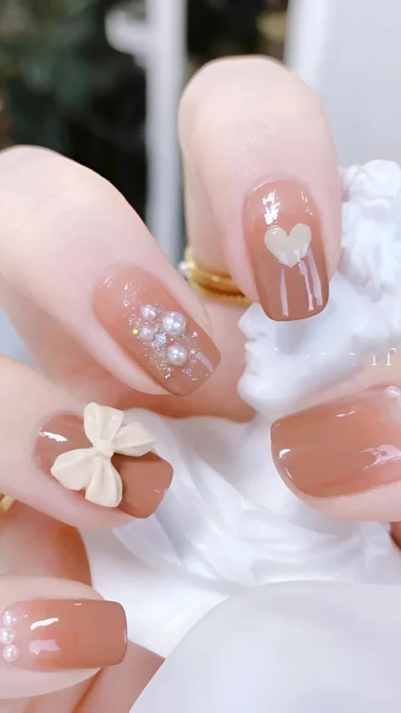 Cute nail design on short square nails with a rosy nude base, featuring varied accents: pearl cluster, 3D cream bow, painted heart, and pearl tip details.