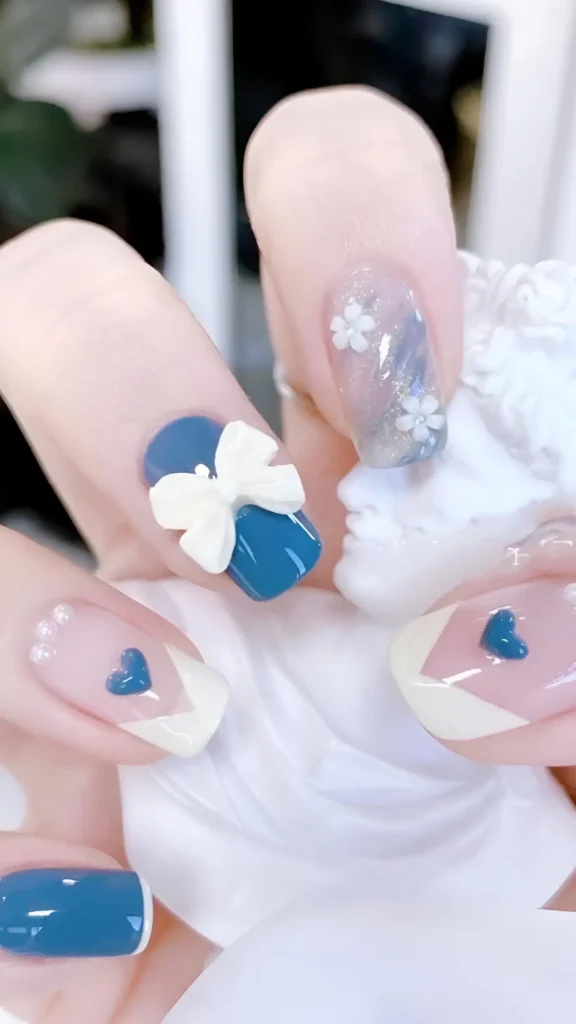 Cute nail design set featuring dusty teal blue and white; includes 3D bow, 3D flowers on cloudy base, V-shaped French tips with dimensional hearts, and pearl accents on short square nails.