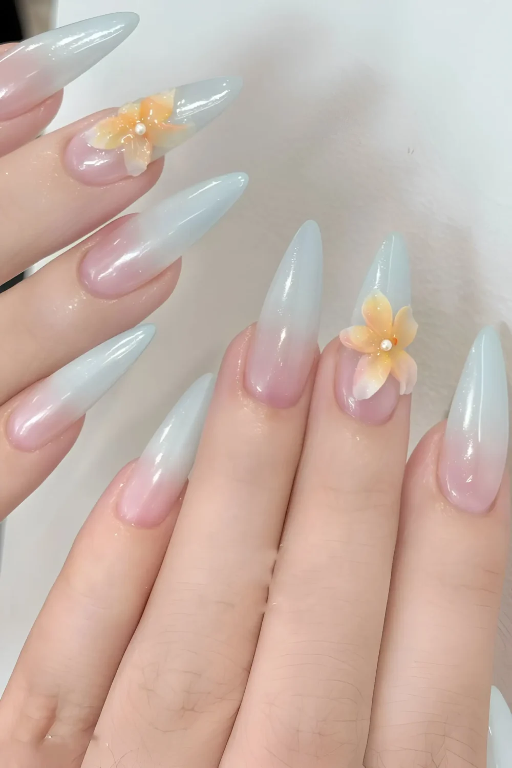 Cute long stiletto nails with pink to pale blue ombre gradient and 3D peach flower charms with pearl centers.