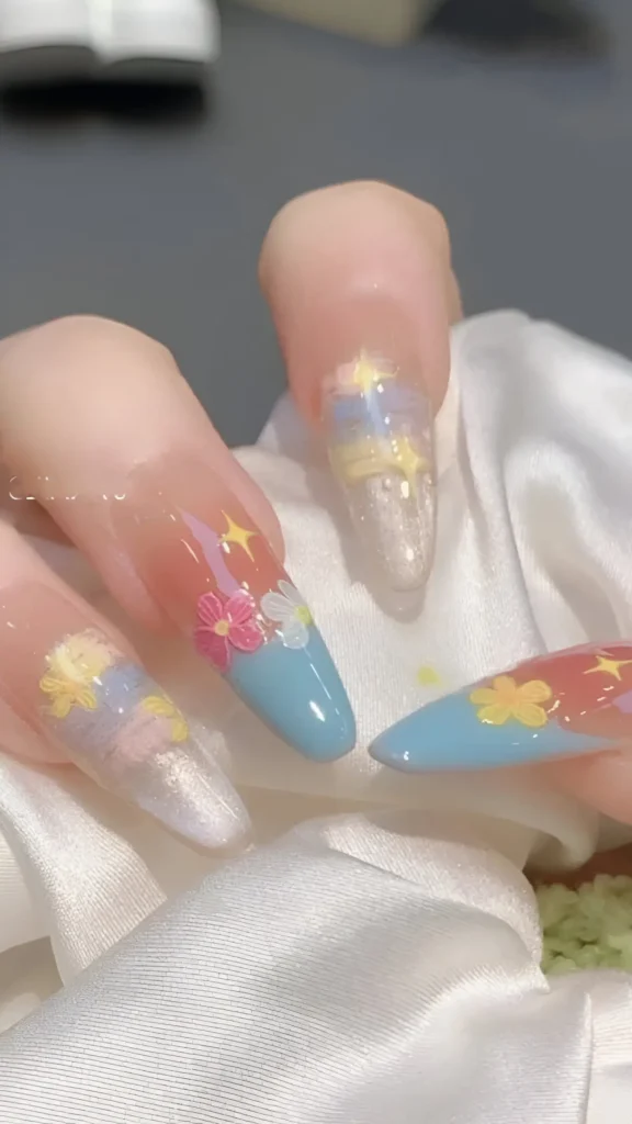 Cute celestial-themed nail design on long almond nails featuring sky blue tips, iridescent shimmer, pastel floral accents (pink, yellow), and yellow starburst details.