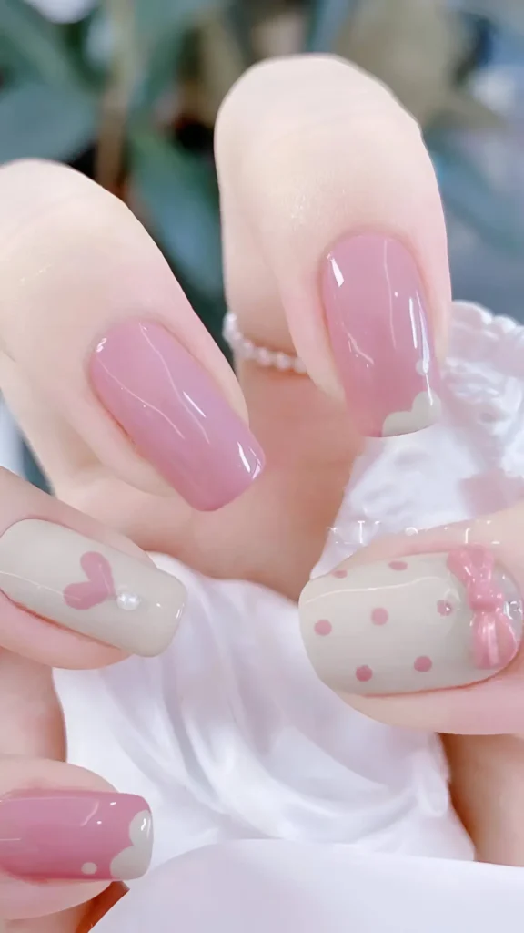 Cute nail design featuring pink nails with white cloud tips, and beige nails with pink polka dots, 3D pink bow, and pink heart motif; medium square shape.