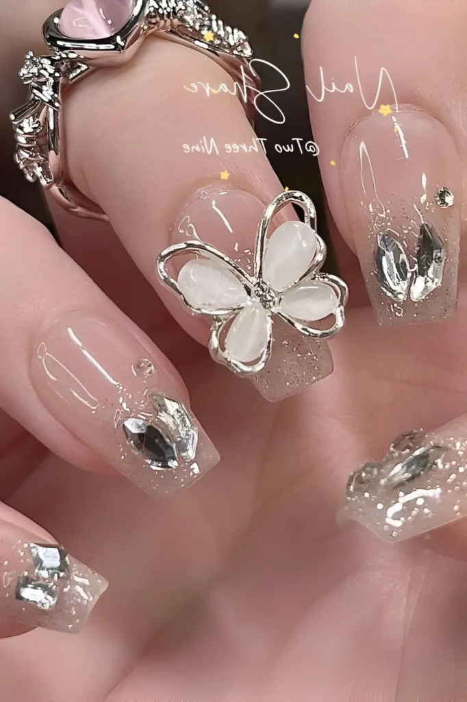Luxury 3D nail art on square nails with glitter tips, featuring a large silver and pearlescent white butterfly charm and scattered clear faceted rhinestones.
