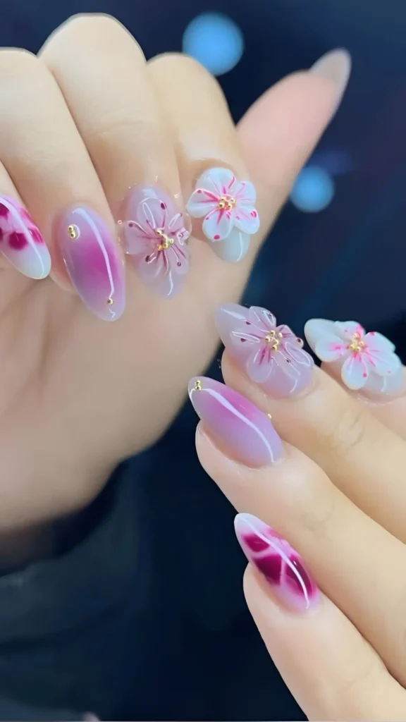Cute nail design on almond nails featuring pink gradients, abstract magenta/white tips, and large 3D pink and white floral appliques (sakura style) with gold bead centers.