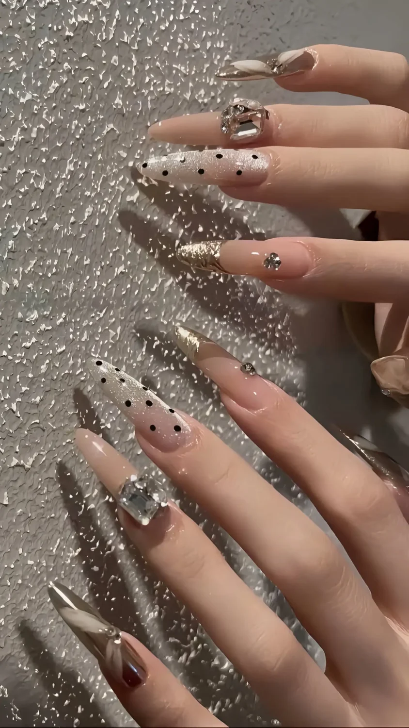 Cute long stiletto nails with mixed designs: black polka dots, metallic accents, and large rectangular crystal gems.