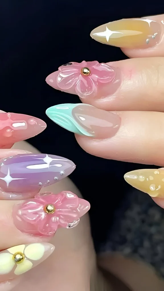 Cute eclectic nail design on long stiletto nails featuring pastel mix (yellow, pink, lilac, green), jelly & iridescent finishes, 3D pink flowers, white starbursts, bead accents, and abstract tip.