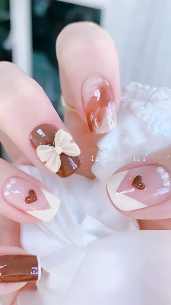 Cute nail design featuring chocolate brown and cream; includes 3D bow, dimensional hearts with V-tips, pearl accents, brown marble effect, and solid brown nails on oval/square shape.