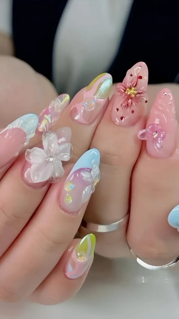 Cute fairycore nail design on long almond nails; sheer base with iridescent/metallic swirls (pink, blue, lime) and prominent 3D floral appliques (pink, white, clear) with accents.