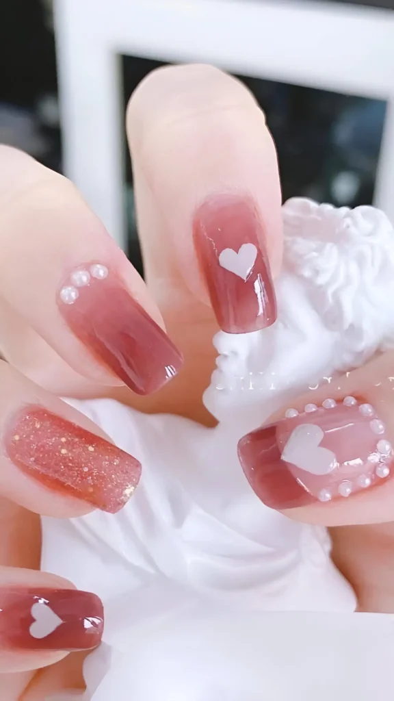 Cute nail design with rosy-brown jelly finish featuring white heart accents, pearl details (some outlining hearts), and fine glitter on square nails.
