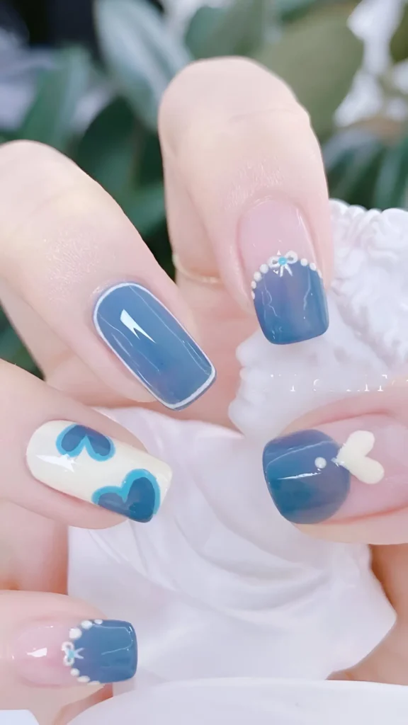 Cute nail design featuring dusty blue French tips with bow/pearl details, solid blue outline nail, blue heart pattern on white, and 3D white heart accent; square shape.