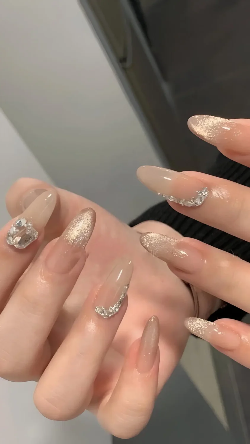 Cute long almond nails with sheer base, champagne gold glitter and cat-eye effects, and clear crystal gem accents.