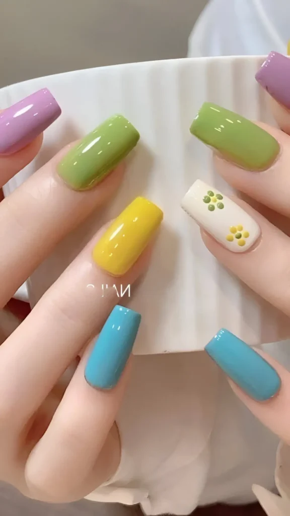 Cute nail design featuring glossy solid colors (lilac, olive, yellow, turquoise) with one white accent nail showing a dot-art flower motif; square shape.