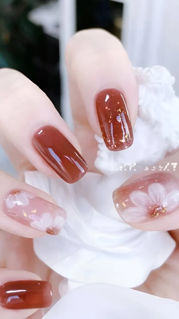 Cute nail design featuring reddish-brown nails with gold foil, and sheer base nails with white floral painting (using brown centers); square shape.