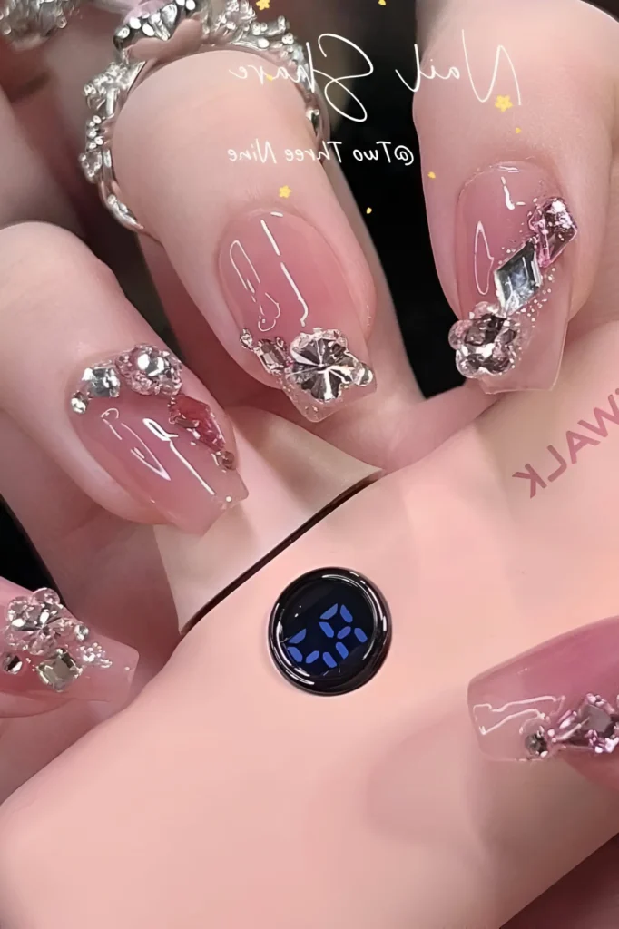 Luxury rhinestone nail design on sheer pink coffin nails, featuring ornate cuticle clusters composed of metallic floral/star studs and mixed clear and champagne crystals.