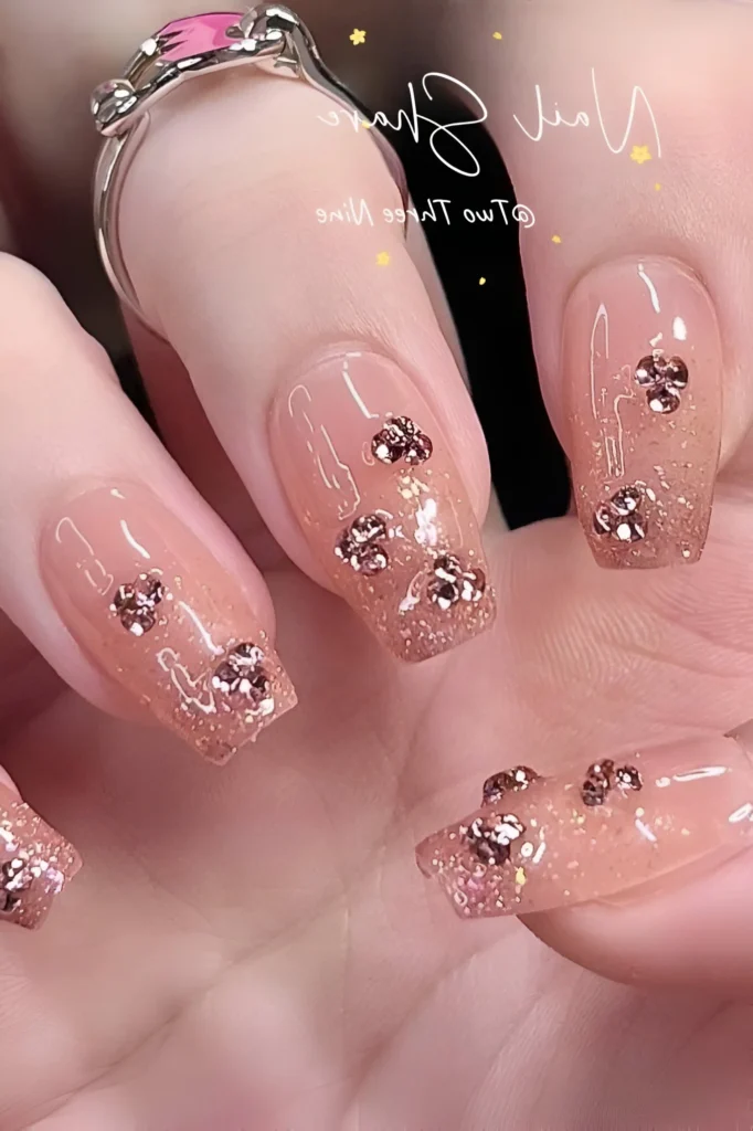 Luxury nail design on sheer pink square nails with a rose gold glitter ombre, embellished with scattered clusters of small metallic floral/clover studs.