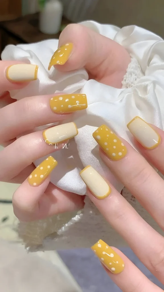 Cute nail design alternating between sunny yellow with white polka dots and creamy white with yellow French tips; square shape.