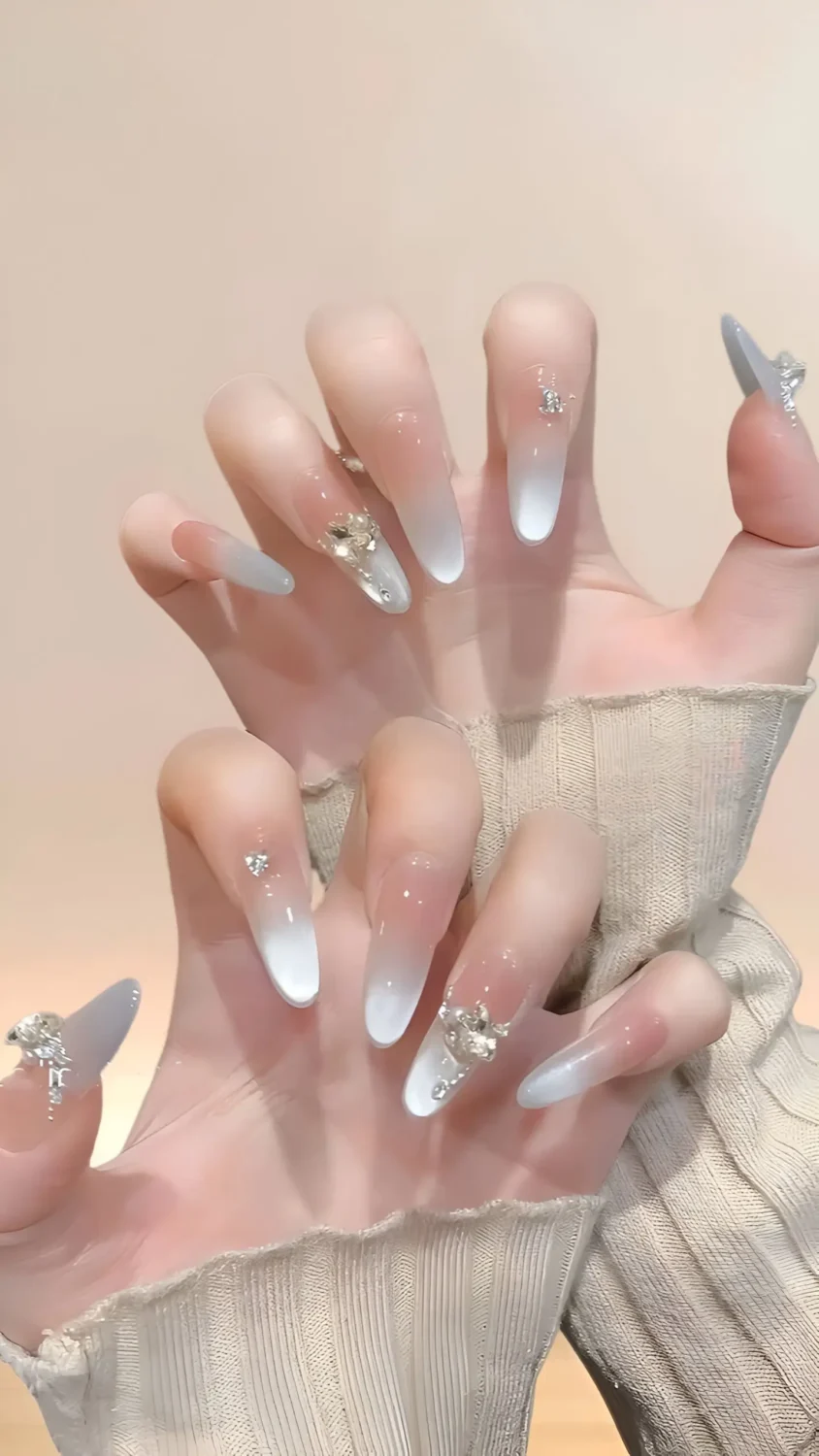 Cute long almond nails with soft nude-to-white ombre base and varied clear crystal gem accents near cuticles.