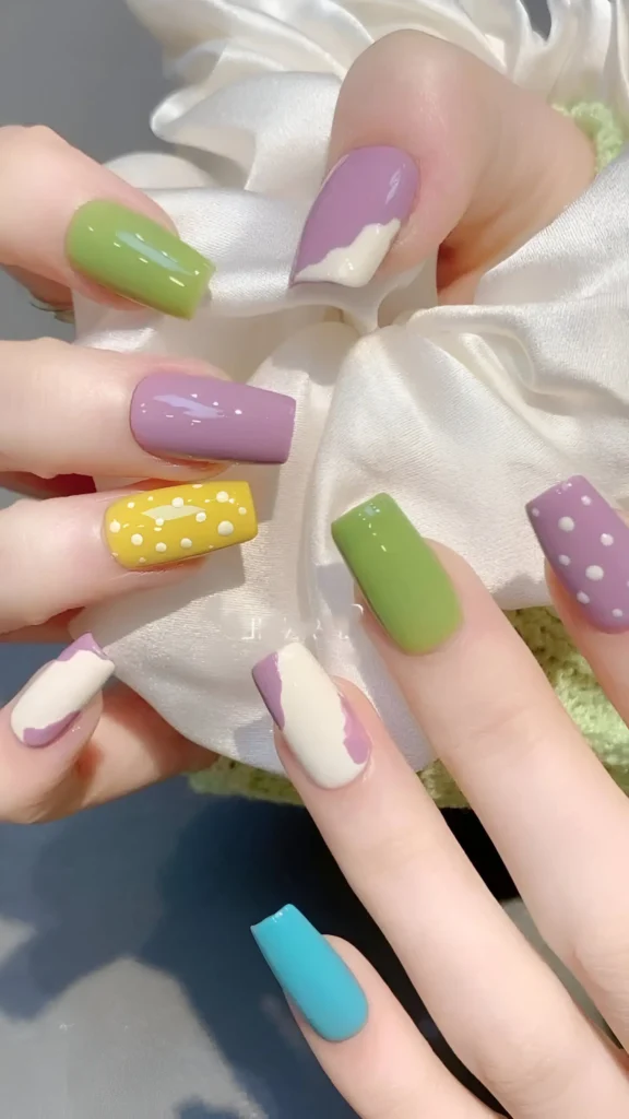 Cute nail design mixing bright solid colors (green, blue) with patterned pastel nails (yellow/lilac polka dots, white with abstract lavender waves); square shape.