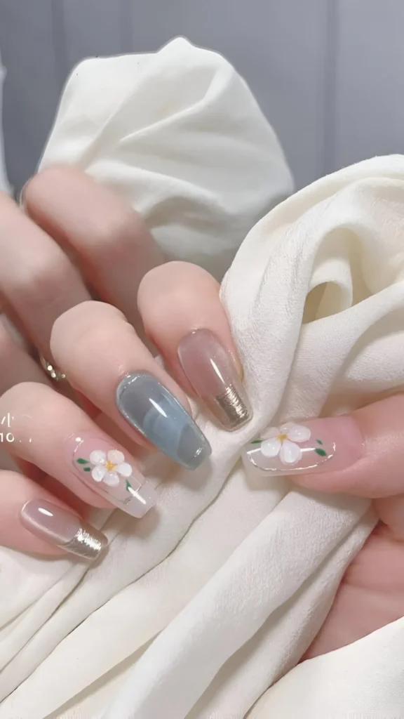 Cute nail design mixing white florals on sheer base, dusty blue-grey marble effect, and metallic gold/bronze tips; coffin/square shape.