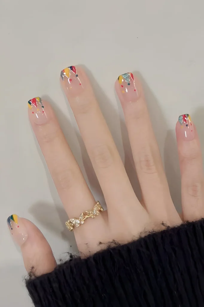 Cute nail design on sheer base featuring abstract confetti/splatter French tips with bright colors (red, yellow, blue) and silver glitter; square shape.