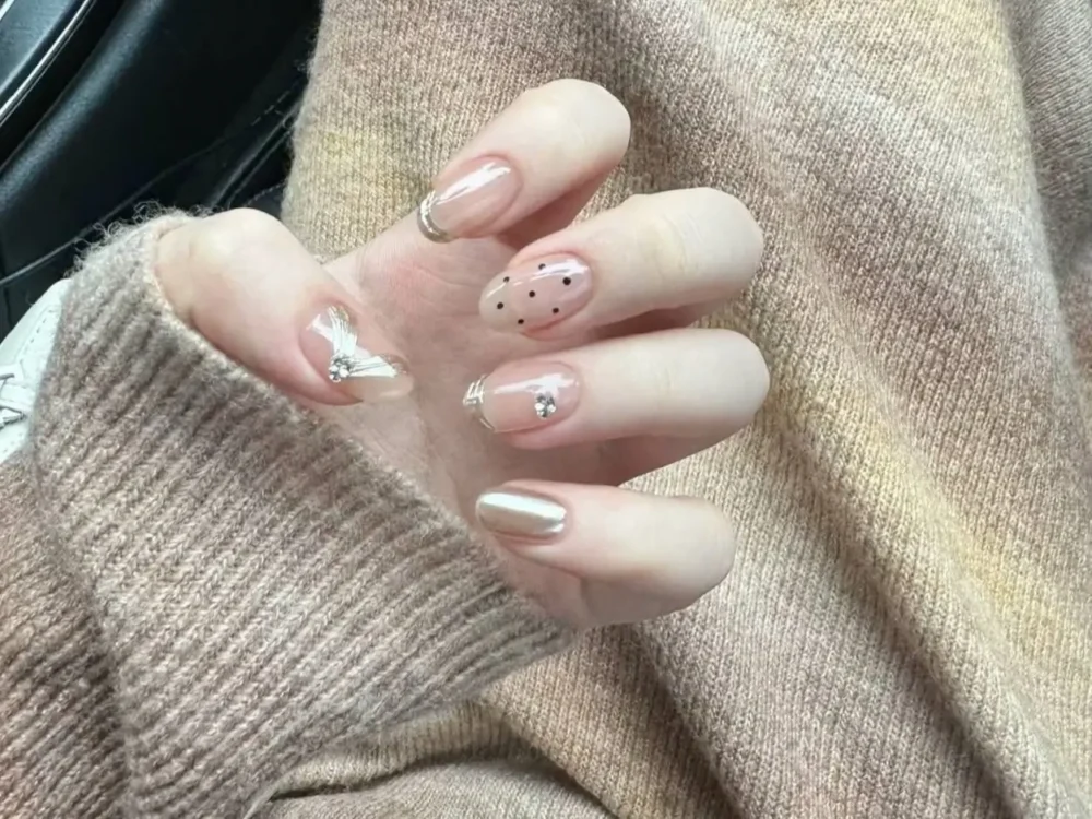 Short oval nails with a charming medley of dainty designs including pale gold chrome, tiny black polka dots, fine line art, and a 3D bow accent.