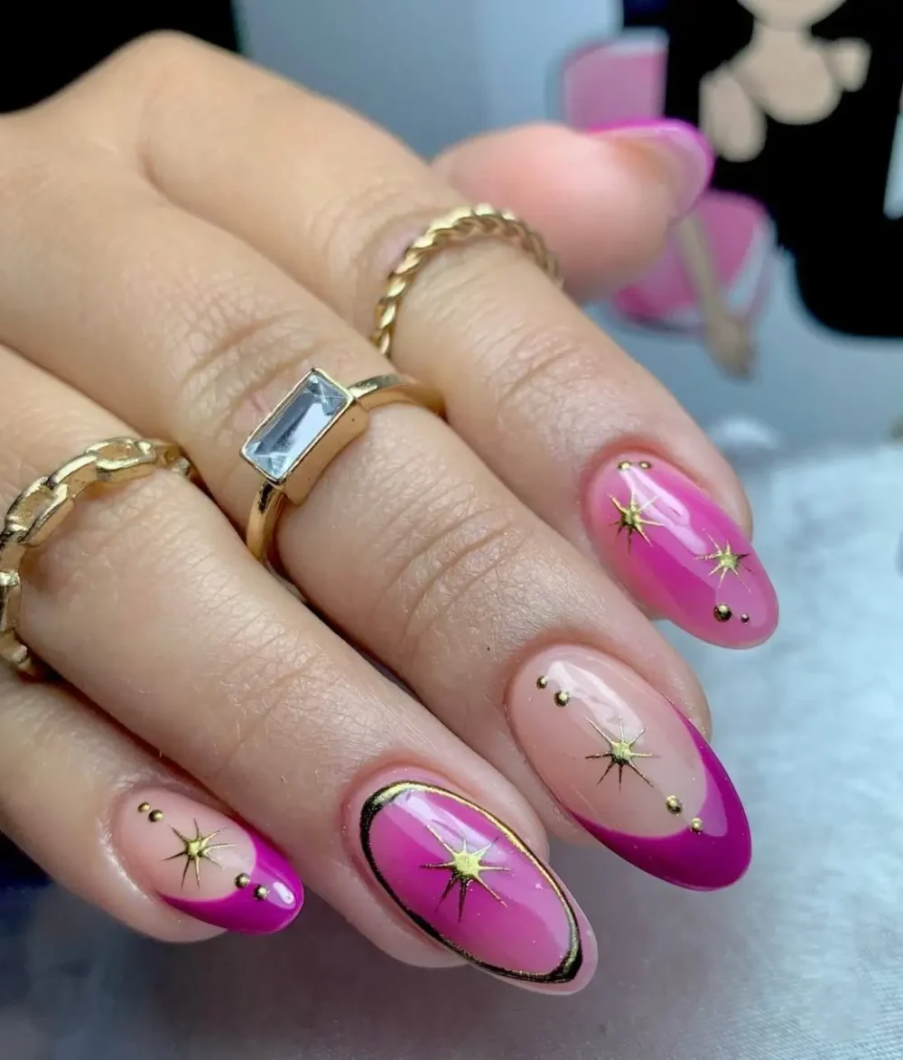 Oval-shaped nails with vibrant fuchsia pink half-moon color blocks on a sheer base, adorned with gold starbursts and tiny stud details.