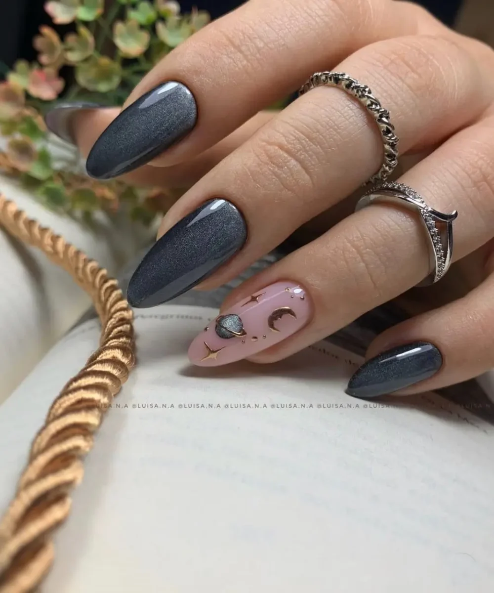 Almond-shaped nails in a shimmery charcoal grey, with a contrasting pink accent nail adorned with delicate gold celestial charms like planets and moons.