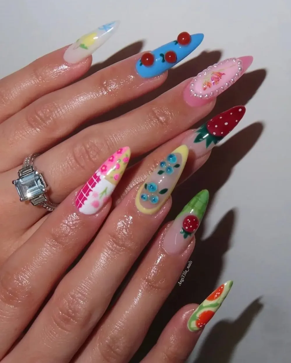 A maximalist manicure on long stiletto nails with mixed art including gingham, 3D strawberries, and florals.