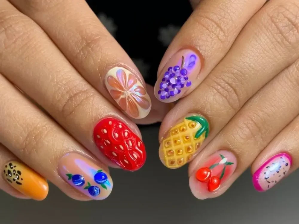 A close-up of a colorful 3D cute fruit nail art medley, featuring a sculptural strawberry, pineapple, grapes, and cherries on oval-shaped nails.